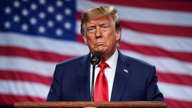 [Trump] at podium, serious expression, American flag background.